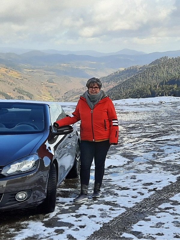 Ute neben der Fahrertür ihres braunen Cabrios in verschneiter Landschaft in den Bergen